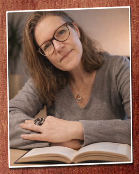 Eva Gitta sitzt an einem Schreibtisch, den Kopf leicht geneigt mit Blick in die Kamera, freundlich, vor ihr auf dem Tisch liegt ein aufgeschlagenes Buch oder Archiv